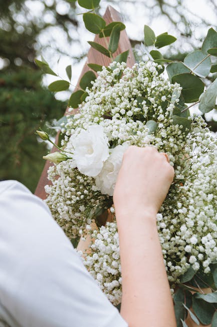 A person’s hand holding a large floral bouquet consisting of white Gypsophila (baby's breath), white Lisianthus, and green eucalyptus leaves, wrapped in brown kraft paper with a pink ribbon, set out
