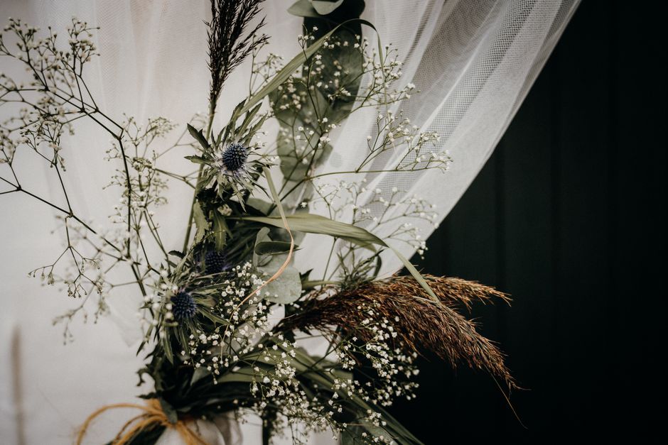 A bouquet arrangement featuring a combination of wildflowers and foliage, including delicate white baby's breath, spiky blue thistles, and wispy brown and green grasses, tied together with a natural j