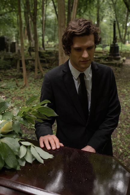A young man with curly brown hair dressed in a formal black suit and white shirt, standing outdoors in a lush, green wooded area during daytime. He appears somber, with his hands resting gently on a p