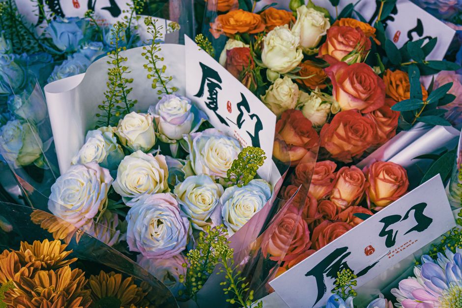 A vibrant arrangement of fresh flowers featuring cream and blush-hued roses, alongside delicate yellow-green accents, nestled in white floral wrapping paper with subtle pink edges. The bouquet is set 