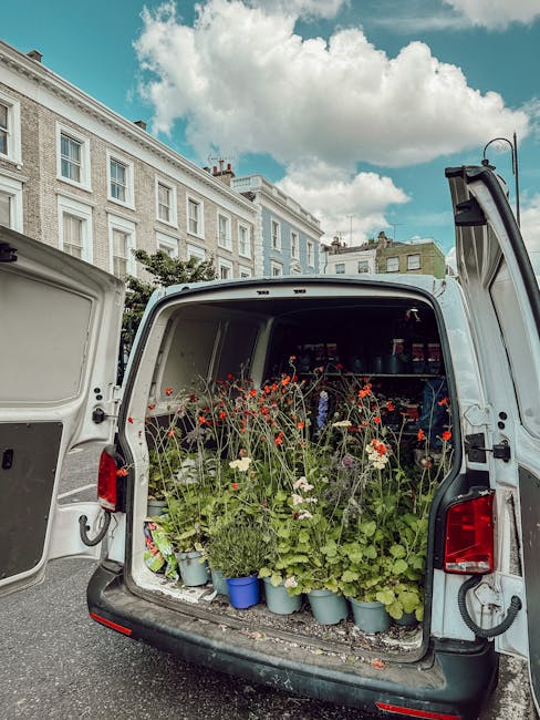 Open rear doors of a white van filled with various potted plants and blooming flowers, including red poppies, white daisies, and green foliage, arranged in blue and grey pots on a gravel surface. In t