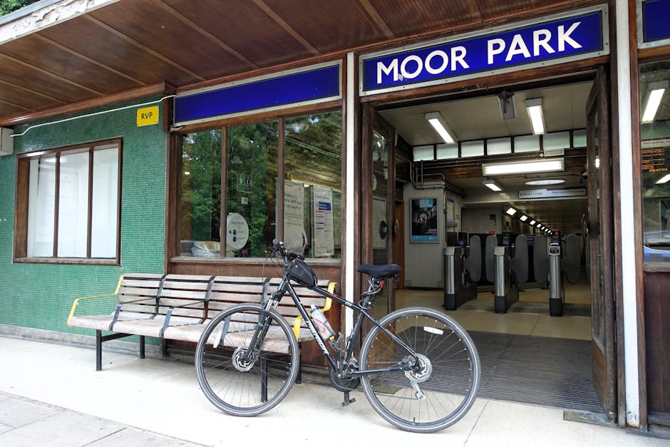 A black bicycle parked in front of the entrance to Moor Park station, featuring a wooden bench with metal armrests beside it. The station has a green tiled exterior wall with a large glass window disp