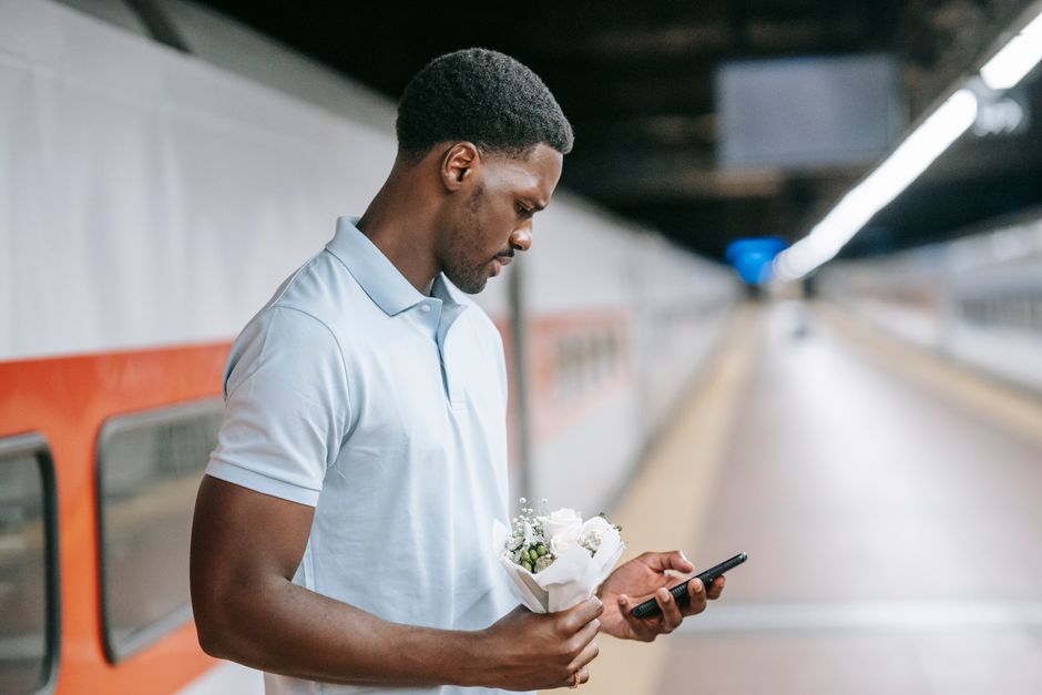 A young man at Honor Oak Park station stands on the platform holding a small bouquet of fresh white flowers, including roses and baby's breath, wrapped in white tissue paper. He is wearing a light blu