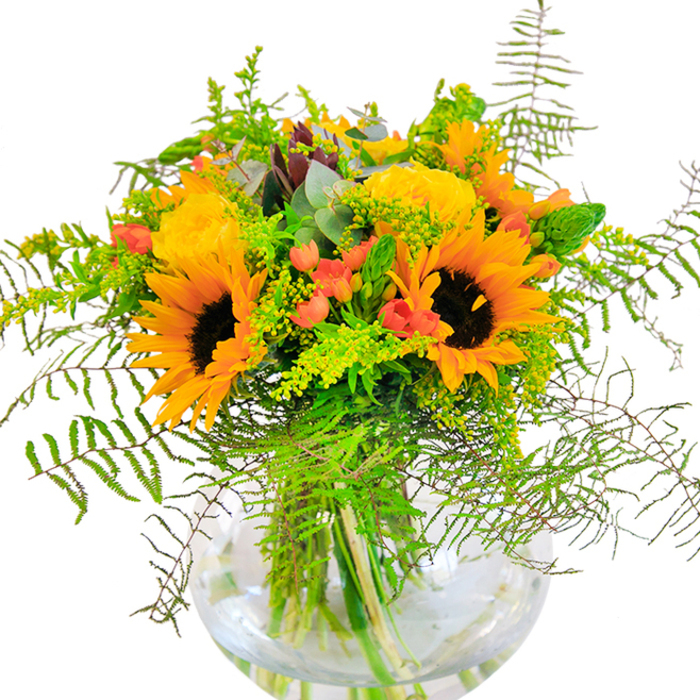 On first glance, this arrangement is an exuberant ode to sunshine and the abundant greenery that flourishes throughout Honor Oak. At the center, vibrant sunflowers beam outward, their golden petals and rich brown centers capturing the vitality of a sunny afternoon spent walking near One Tree Hill. Alongside them, plush yellow garden roses spiral open with old-world flair, filling the bouquet with layers of soft, tactile petals and a hint of their delicate fragrance. Glossy red Hypericum berries are artfully interwoven between the larger blooms, their vibrant scarlet hue offering both contrast and a playful spirit. The interplay of deep green fern fronds, light sprigs of filler-potentially locally sourced solidago or goldenrod-and the gentle blue-green of eucalyptus leaves creates a rich, textural tapestry reminiscent of wild woodlands at the edge of Honor Oak. The arrangement is anchored in a crystal-clear, round glass vase that allows each stem to shine, giving a sense of structure and freshness that celebrates the artistry of Flowers Honor Oak's team. Whether gracing a table for a milestone celebration or acting as a cheerful centerpiece for gatherings around Brockley, the bouquet radiates positivity, creativity, and a uniquely local sense of vibrant life.